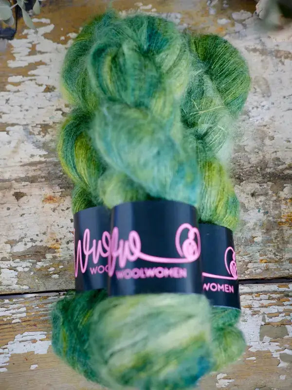 A skein of variegated green and yellow WoolWomen Fluff Mohair with a black paper label wrapped around the center.