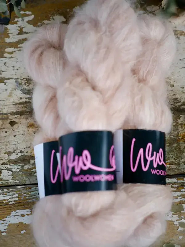 A skein of pale pink fuzzy WoolWomen Fluff Mohair with a black paper label wrapped around the center.