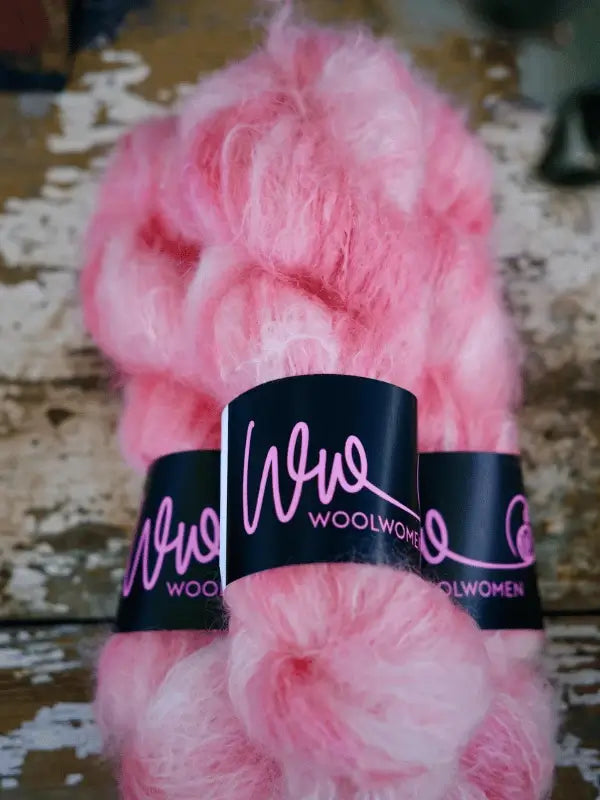 A close-up of two skeins of fluffy, pink WoolWomen Fluff Mohair yarn with black and pink tags, resting on a rustic wooden surface.