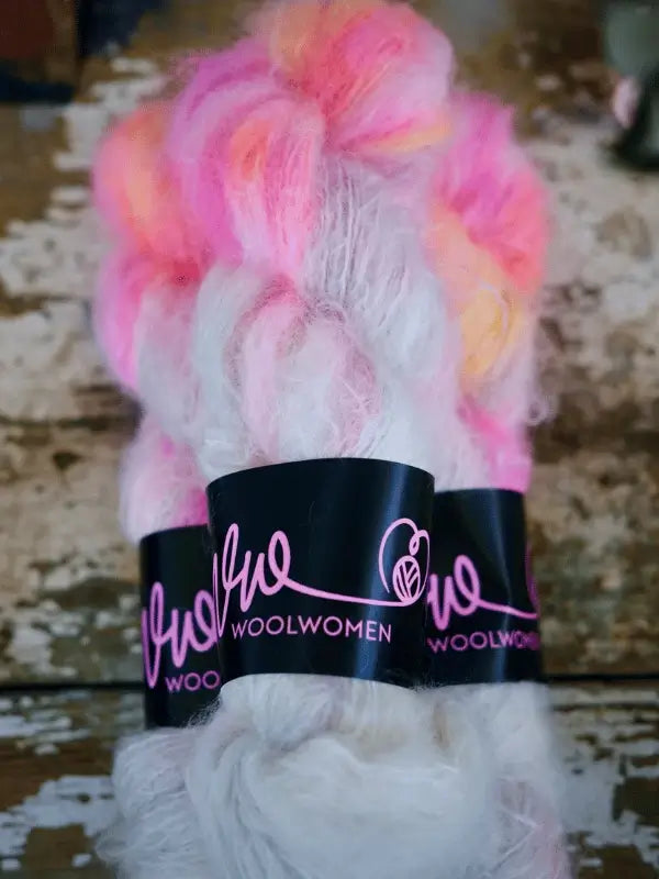 Skeins of WoolWomen Fluff Mohair yarn in pink, white, and orange shades are wrapped in black labels with pink cursive "WoolWomen" text, arranged on a rustic wooden surface.