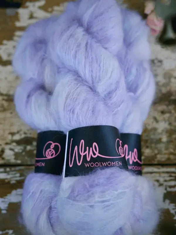 Two skeins of WoolWomen Fluff Mohair in soft, light purple with a fluffy texture, wrapped in black labels with pink script, rest on a rustic wooden surface.
