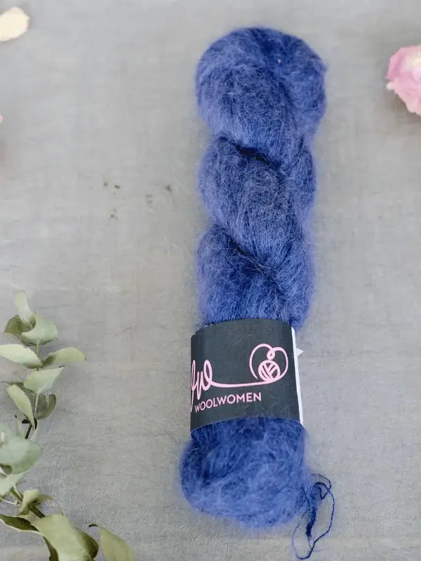 A skein of deep blue WoolWomen Silk Mohair yarn with a black "woolwomen" label rests on a gray surface, accented by small plant leaves and a pink flower in the corners.