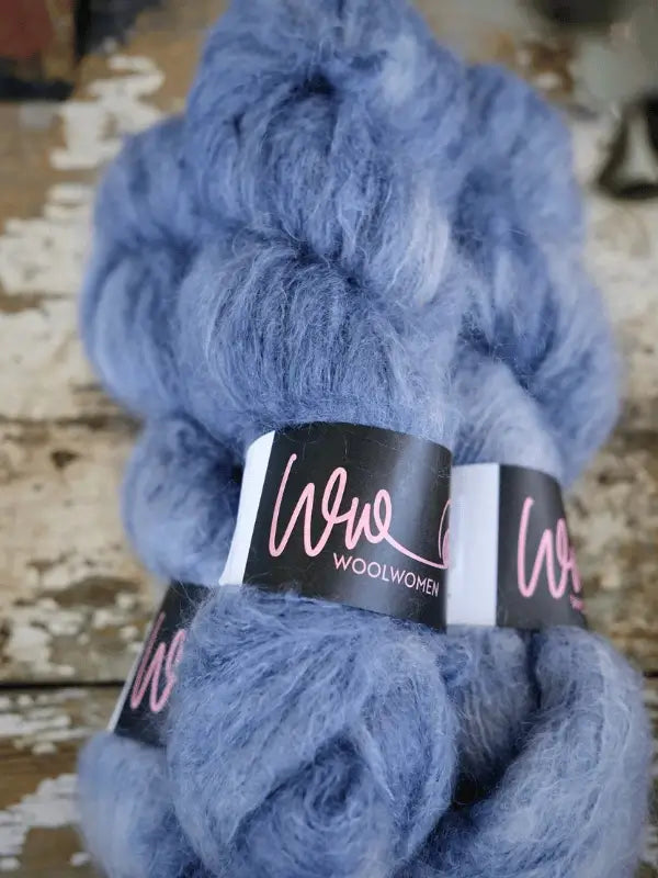 Two skeins of fluffy, light blue WoolWomen Fluff Mohair DK-weight yarn with pink and white labels rest on a weathered wooden surface.
