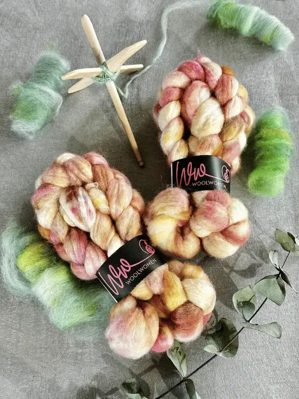 Two skeins of WoolWomen Kehruuvilla BFL Käsinvärjätyt with labels, a wooden drop spindle spun with green Bluefaced Leicester wool, two small bundles of carded green fiber, and eucalyptus on gray—perfect for kehräämiseen.