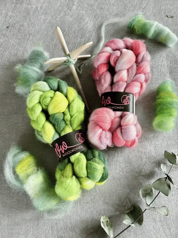 Two bundles of WoolWomen Kehruuvilla BFL Käsinvärjätyt, one green and one pink, rest on gray fabric. Nearby are a wooden spinning spindle, loose wool strands, and a dried eucalyptus sprig.