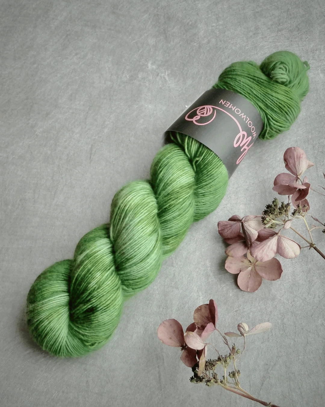 A skein of WoolWomen Merino Single in bright green rests on a gray surface beside dried pinkish flowers. Its label displays a pink logo and text.