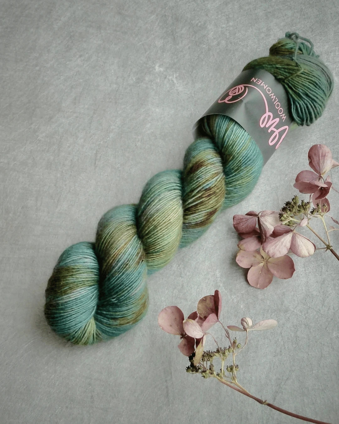 A skein of WoolWomen Merino Single, variegated green and brown yarn with a label, rests on a gray surface beside dried pink hydrangea flowers.