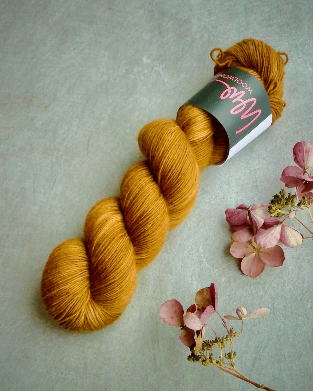 A skein of WoolWomen Merino Single yarn with a label rests on a gray surface next to some dried pink hydrangea flowers.