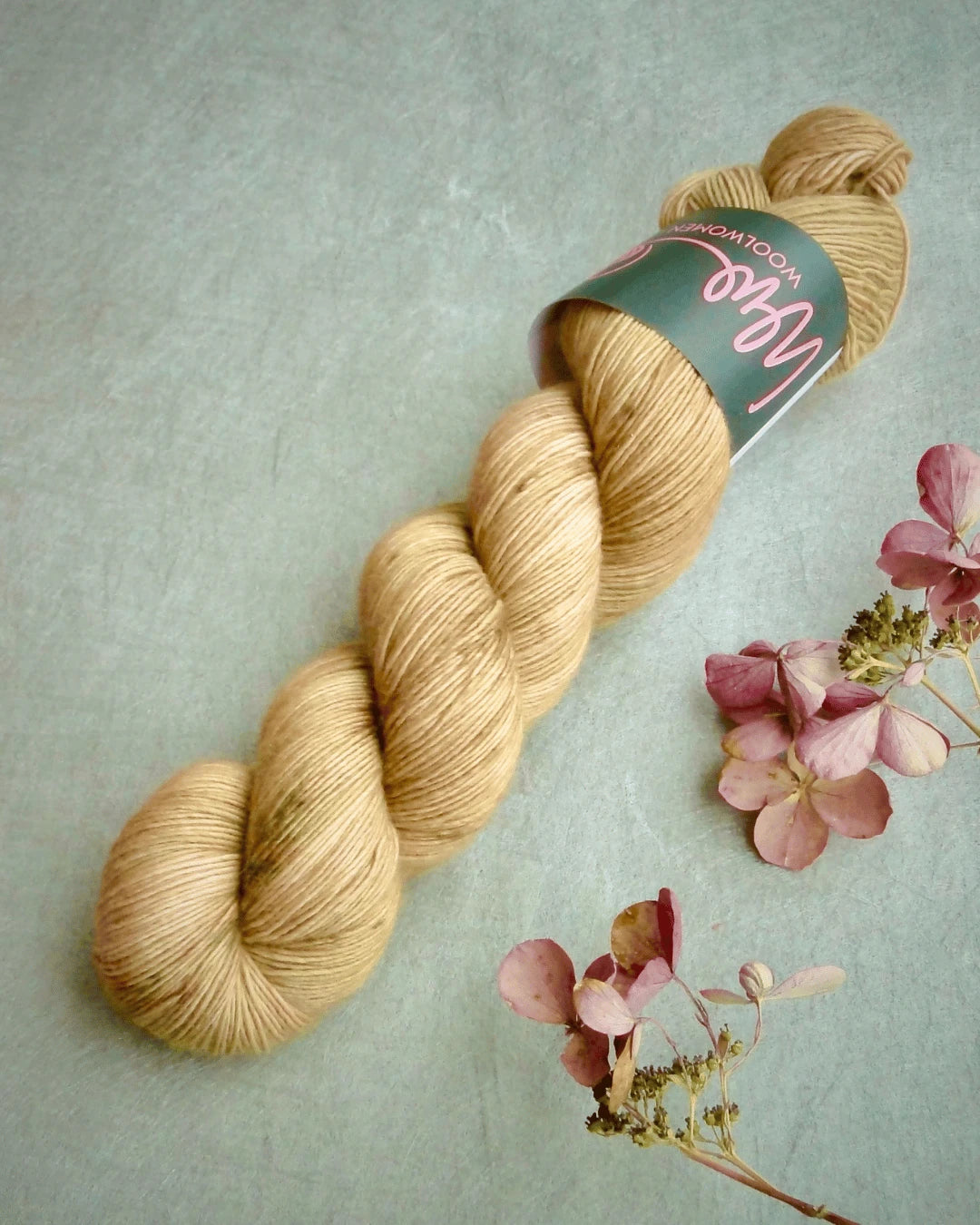 A skein of WoolWomen Merino Single yarn with a label sits on a gray surface beside several pink dried hydrangea flowers.