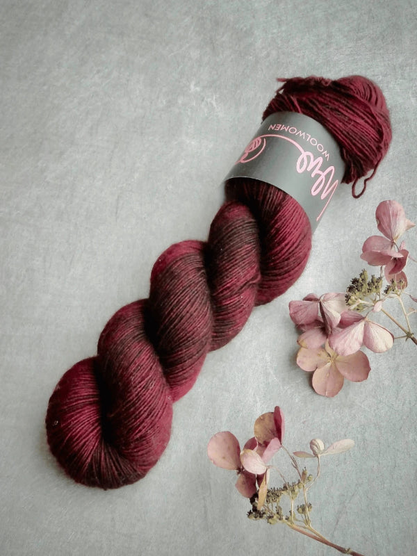 A skein of deep burgundy and dark red WoolWomen Merino Single käsinvärjätty merinovilla yarn rests on a gray textured surface, surrounded by scattered dried pink and beige hydrangea flowers.