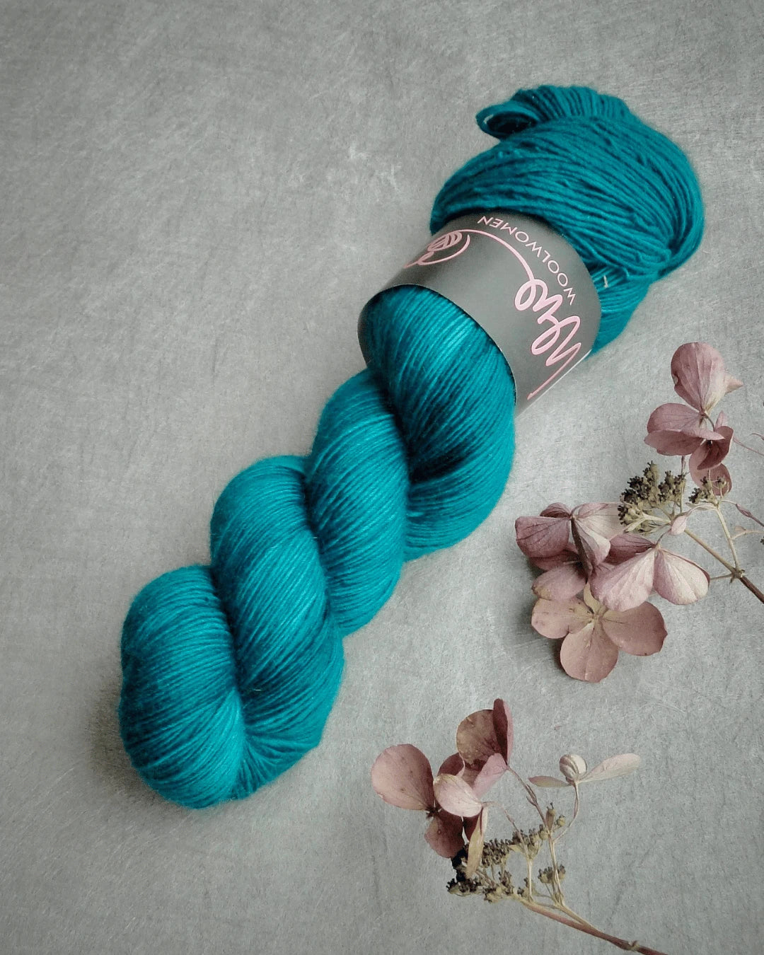 A skein of WoolWomen Merino Single yarn with a label rests on a gray surface next to a few dried pinkish-brown hydrangea flowers.