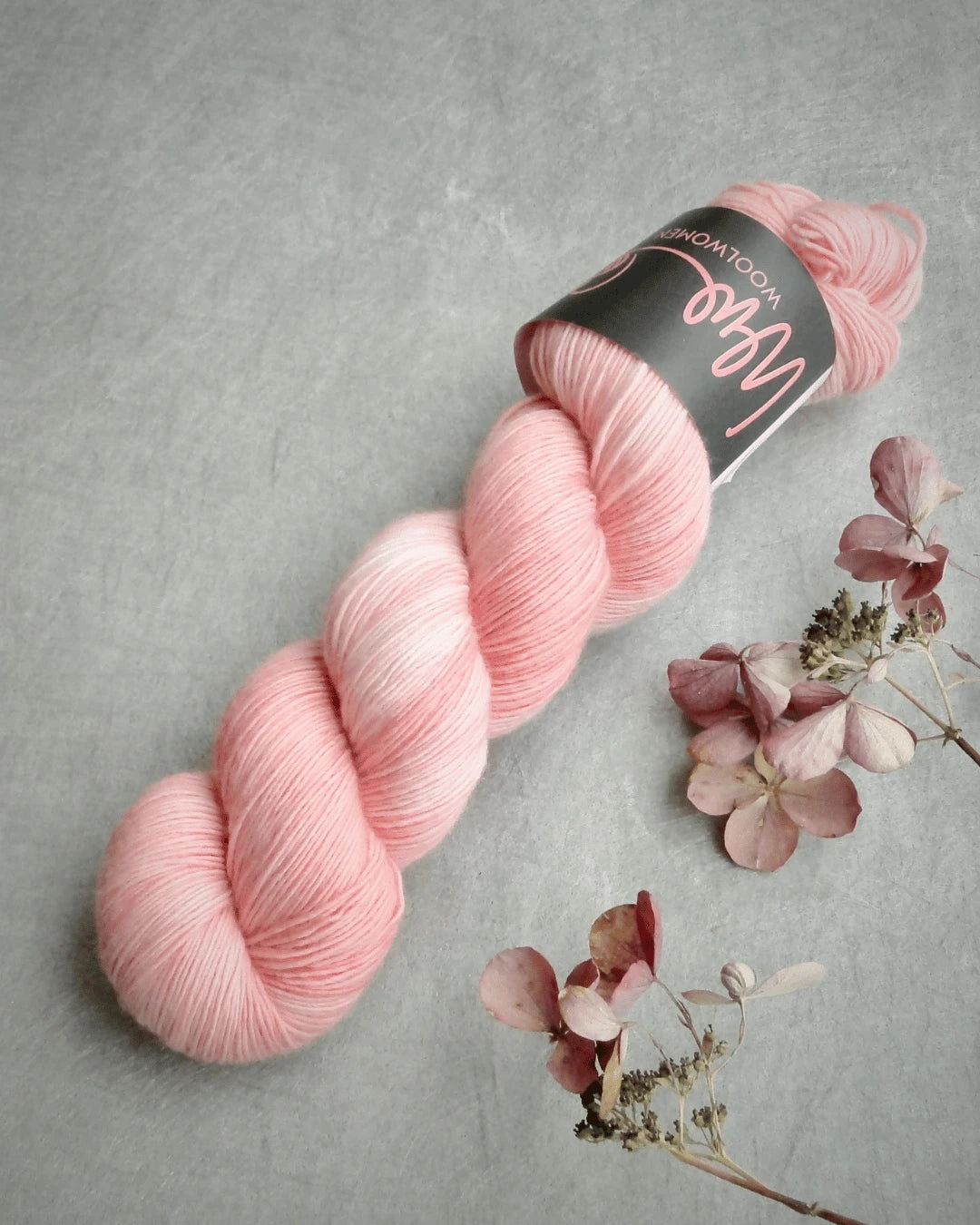 A skein of soft pink WoolWomen Merino Single yarn with a label rests on a gray surface beside dried hydrangea flowers.