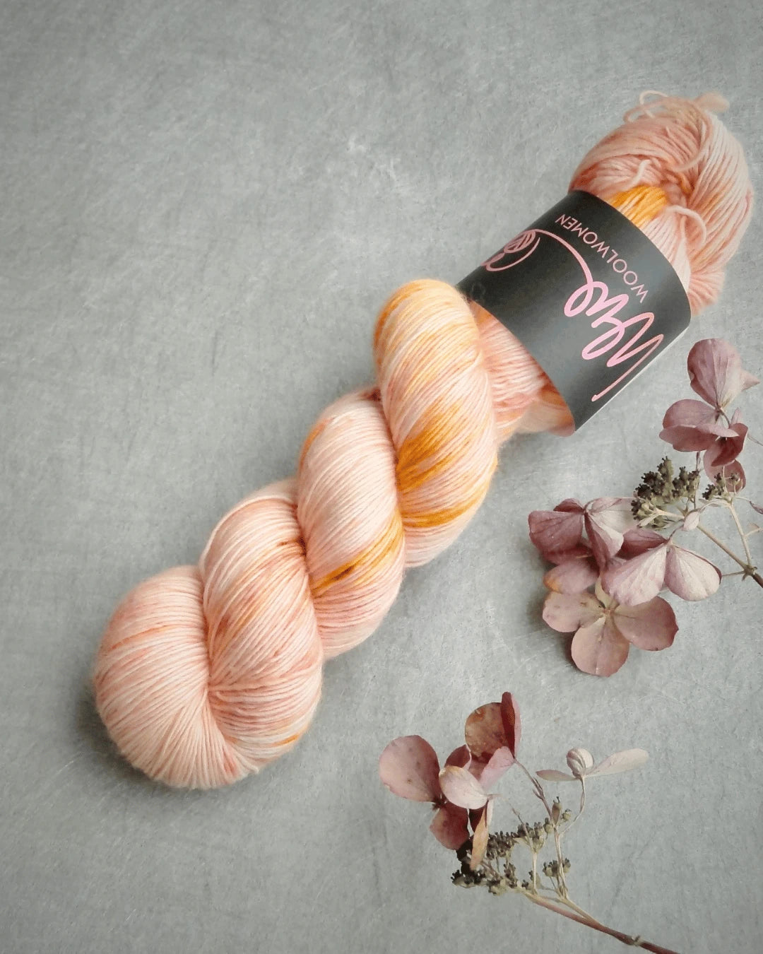 A skein of WoolWomen Merino Single yarn in soft peach and orange tones rests on a light grey surface beside a few dried, muted pink hydrangea flowers.