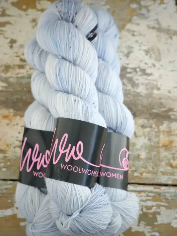 Two skeins of pale blue speckled WoolWomen Merino Sock hand-dyed yarn with black paper labels.