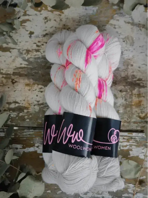 Two hanks of WoolWomen Merino Sock in white with bright pink and orange splatters, tied with black product labels.