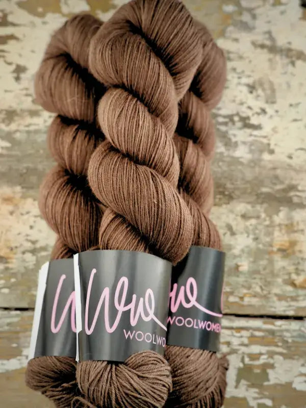 Two twisted hanks of brown WoolWomen Merino Sock yarn with black labels featuring pink script text.