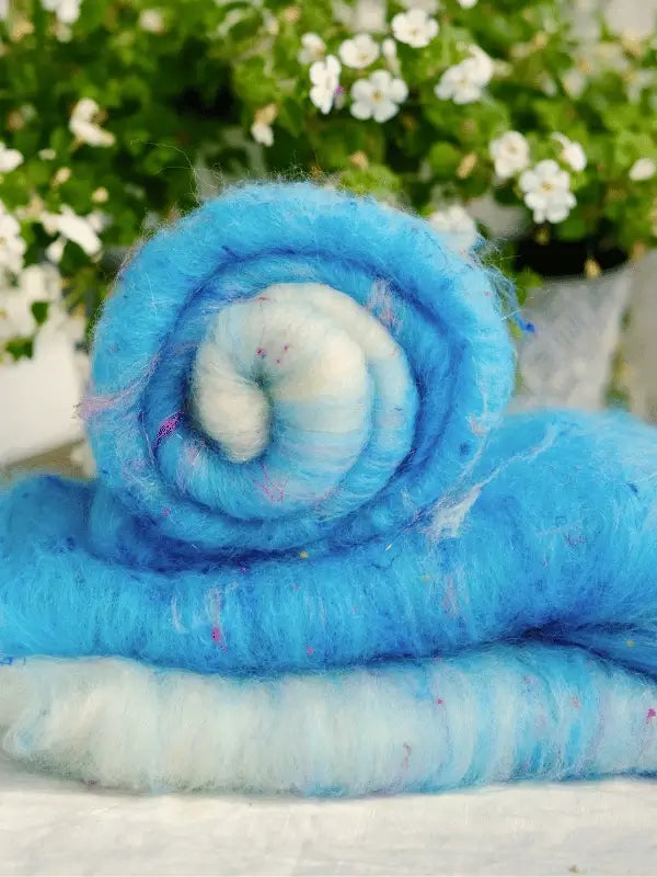 A roll of WoolWomen Art Batt - Corriedale in blue and white rests on a flat surface, ready for spinning, with a blurred background of green foliage and white flowers.