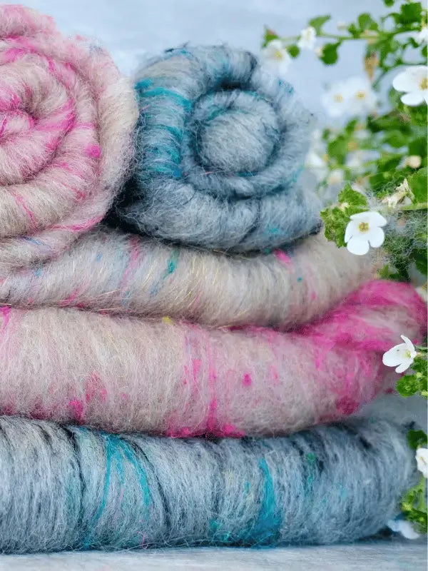 Rolled bundles of WoolWomen Art Batt -BFL in pink, gray, and blue hues are stacked next to small white flowers and green leaves.