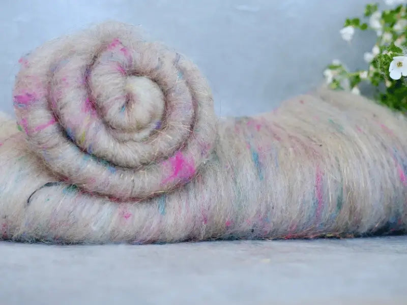 A close-up of a rolled bundle of WoolWomen Art Batt -BFL wool roving with pink and blue specks, lying on a flat surface with small white flowers in the background—ideal for art batt spinning.