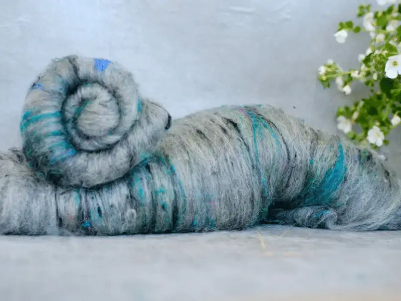 A spiral of WoolWomen Art Batt -BFL, käsinvärjätty villa wool roving with blue and colored flecks, rests on a light surface. Small white flowers and green leaves appear on the right—ideal for kehräys projects.