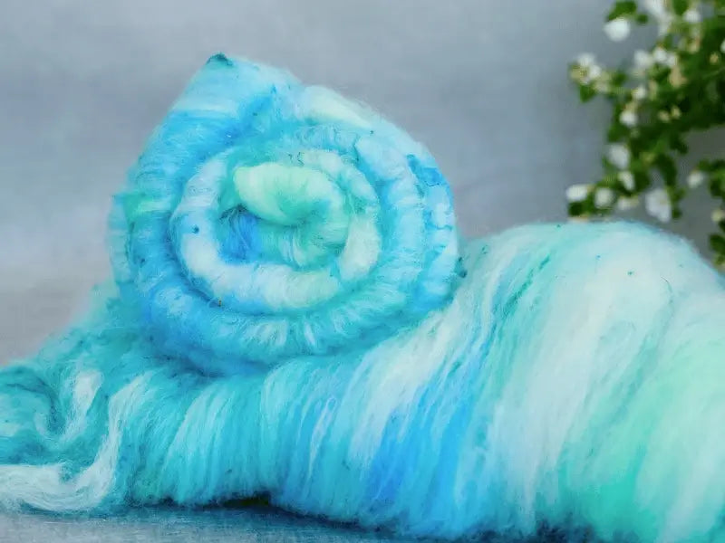 A close-up of WoolWomen Art Batt - Polwarth shows a soft, fluffy roll in light blue and teal with greenish-white fibers. Nearby, small white flowers and green leaves inspire ideas for unique hand-spun yarn.