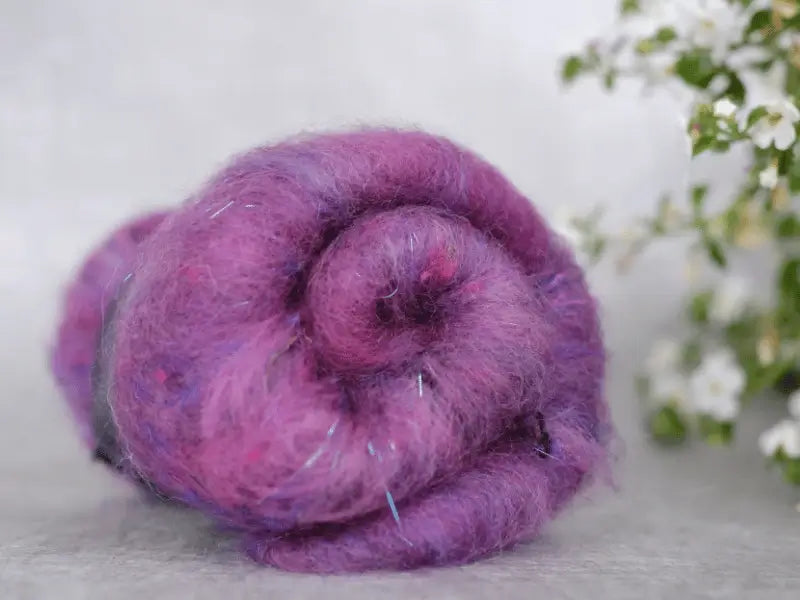 A close-up of a rolled bundle of WoolWomen Art Batt - Suomalainen villa in purple and pink, with subtle blue strands, rests on a light surface. White flowers with green leaves appear blurred in the background on the right.