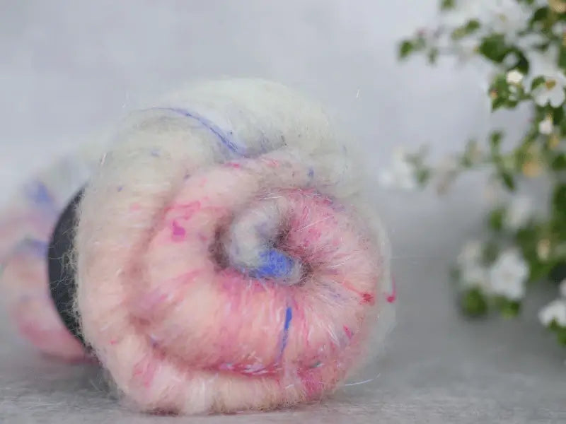 A close-up of a roll of WoolWomen Art Batt -Suomalainen villa in soft white, pink, and light blue shades with small specks. Blurred white flowers and green leaves in the background highlight this beautiful käsinvärjätty villa kehräys.
