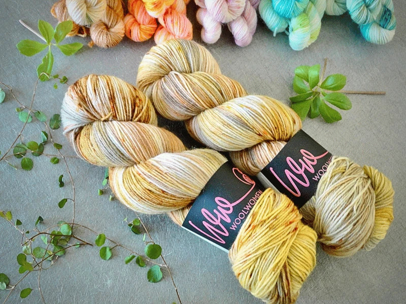 Two skeins of WoolWomen Classic Sock in variegated yellow and brown lie on a gray surface, framed by green leaves and colorful yarns in the background.