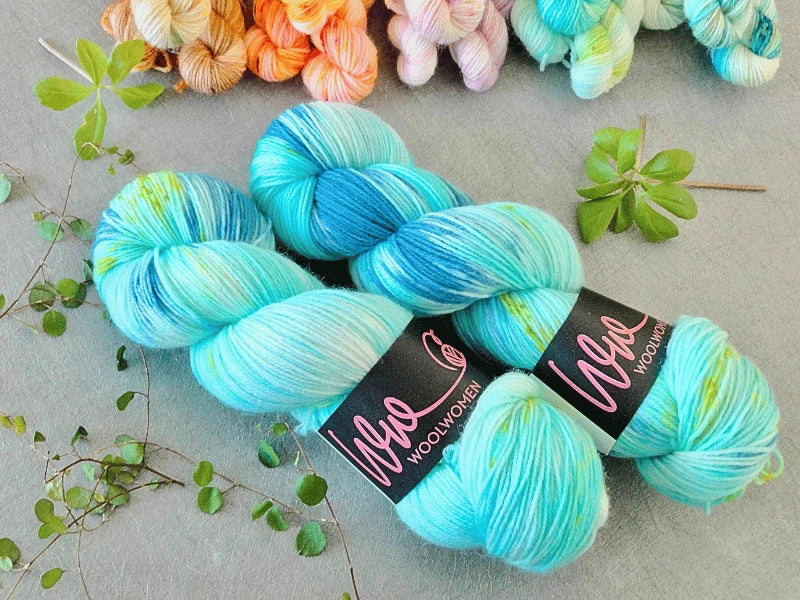 Two skeins of WoolWomen Classic Sock yarn in blue, teal, and white rest on a light gray surface with WoolWomen labels, surrounded by small green leaves and pastel-colored yarns in the background.