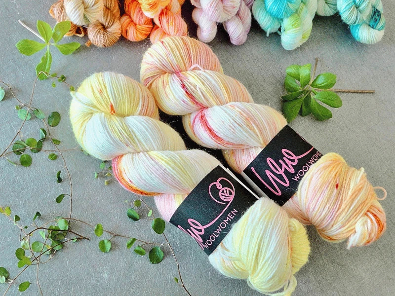 Two skeins of pastel-colored WoolWomen Classic Sock yarn with labels rest on a gray surface. Green leaves and stems are scattered nearby, with additional colorful skeins visible in the background.