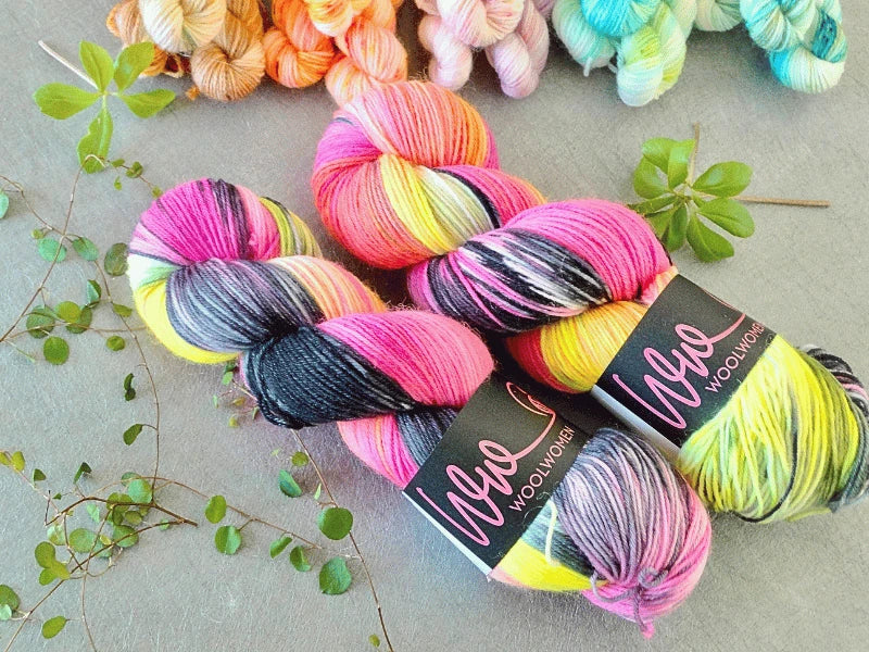 Two skeins of WoolWomen Classic Sock yarn in pink, black, yellow, and white rest on a gray surface with green leaves around them. Extra pastel-colored WoolWomen Classic Sock skeins are partially visible at the top of the image.