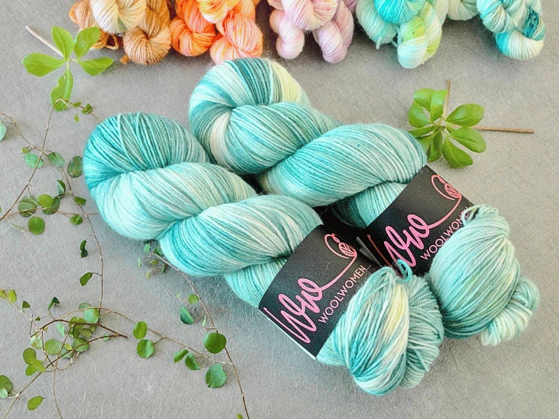 Two skeins of turquoise and white WoolWomen Classic Sock yarn on a light surface, with green leaves and pastel-colored yarn skeins in the background.