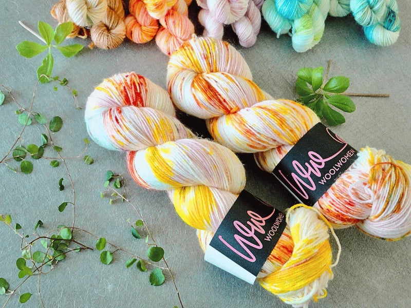 Two skeins of colorful WoolWomen Classic Sock yarn rest on a gray surface, surrounded by green leafy sprigs and additional pastel-hued skeins in the background.