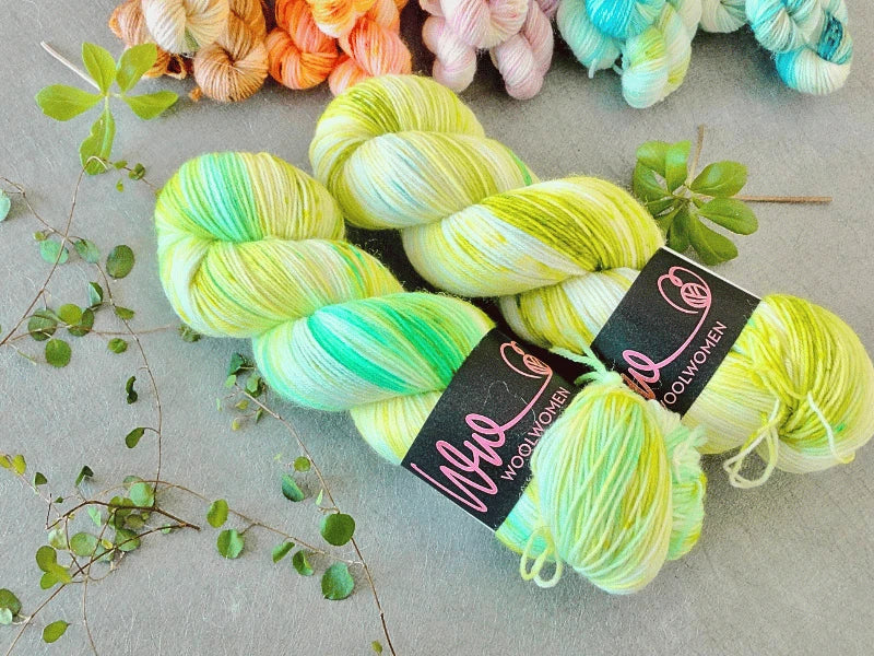 Two skeins of yellow, green, and white WoolWomen Classic Sock yarn lie on a gray surface, surrounded by green leaves and several other colorful yarn skeins in the background.