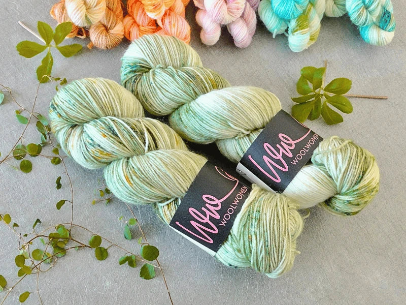 Two skeins of green and white WoolWomen Classic Sock yarn are displayed on a light grey surface with small green leaves. Other colorful skeins are partially visible in the background.