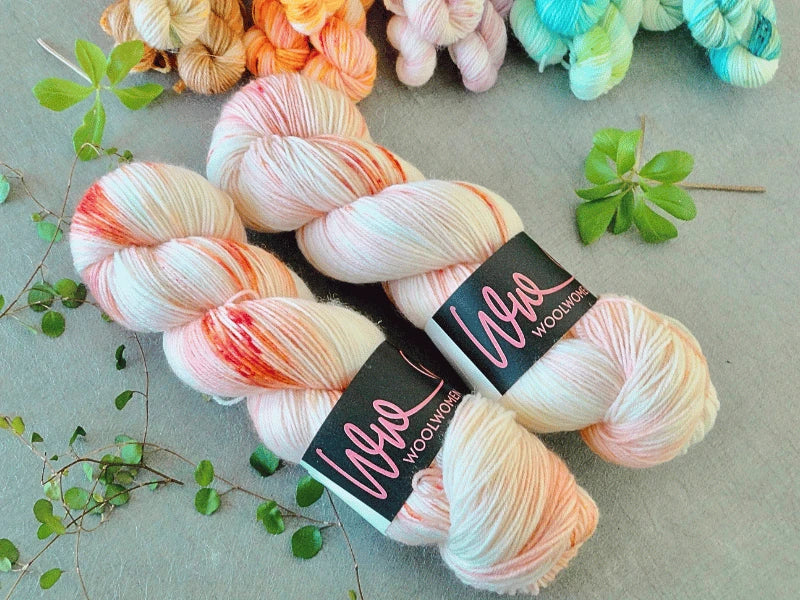 Two skeins of WoolWomen Classic Sock yarn in pale pink and red, labeled “Ula + Lia Woolworks,” are shown on a gray surface with green sprigs and colorful yarn bundles in the background.
