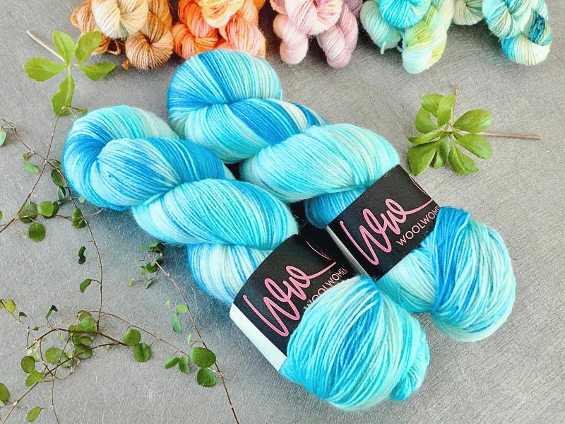 Two skeins of blue and white WoolWomen Classic Sock are displayed on a gray surface, surrounded by green vines and colorful yarn skeins in the background.