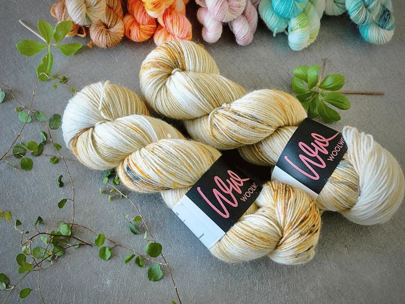 Two skeins of cream-colored, speckled WoolWomen Classic Sock yarn rest on a gray surface with green leafy sprigs. Colorful skeins of yarn are softly blurred in the background.