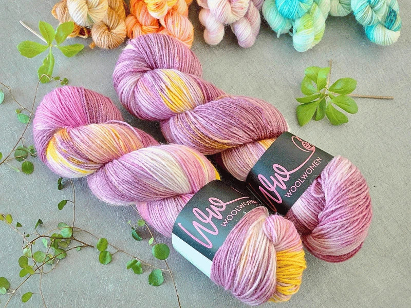 Two skeins of WoolWomen Classic Sock in variegated purple, pink, yellow, and white are displayed on a gray surface with green clover leaves and colorful yarn skeins in the background.