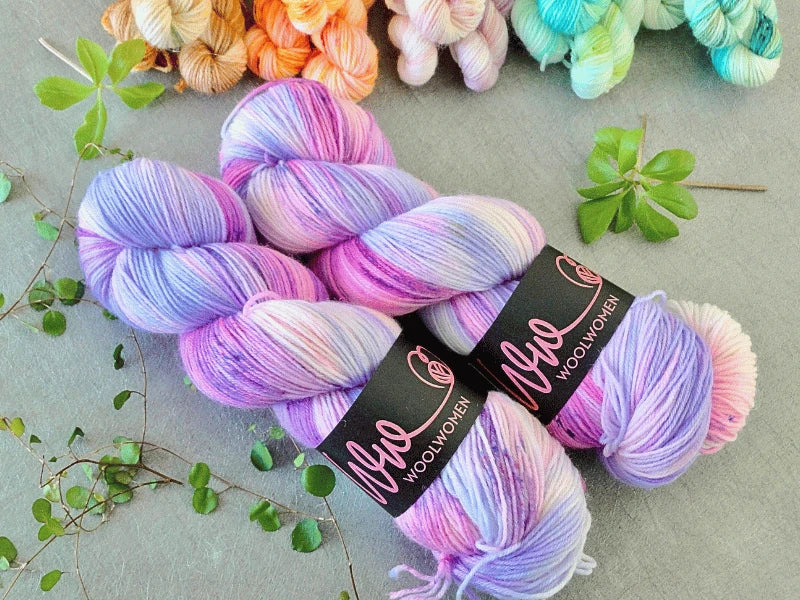 Two skeins of WoolWomen Classic Sock in pastel pink, purple, and blue with Nyo WOOLWOMEN labels rest on a gray surface surrounded by green leaves and other colorful yarns in the background.