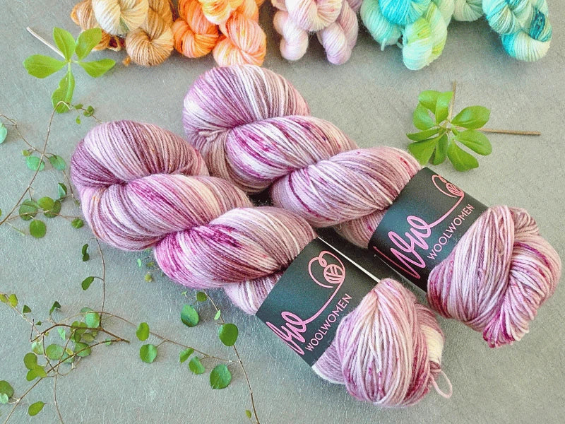 Two skeins of pink and purple variegated WoolWomen Classic Sock yarn lie on a gray surface, surrounded by small green leaves and other colorful yarn skeins in the background.