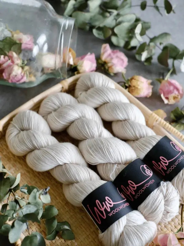 Three skeins of soft, light gray WoolWomen Merino Sock yarn with black labels are displayed in a woven tray, surrounded by dried pink roses and greenery, with a glass jar tipped over in the background.