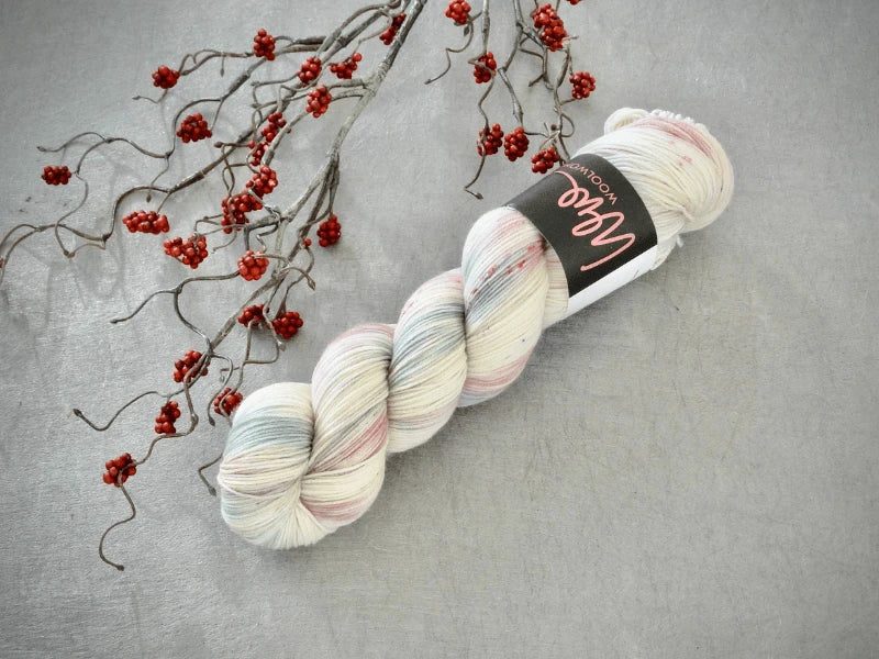 A skein of WoolWomen Merino Sock – Yhden Padan Ihme in soft pastel pink and blue rests on a gray surface beside a branch with small red berries.