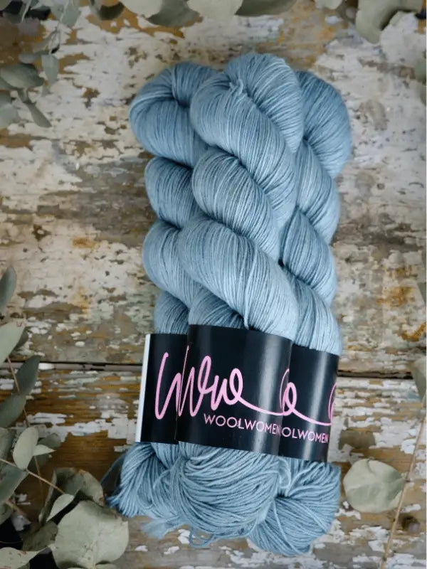 Three skeins of WoolWomen Merino Sock in light blue are neatly stacked on a rustic wooden surface with dried leaves around. Each skein is wrapped in a black label featuring pink handwritten text.