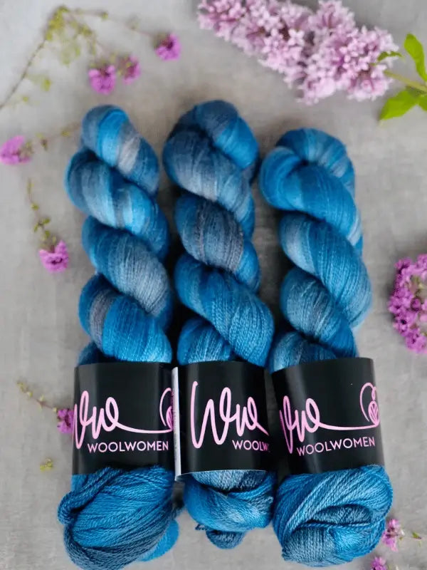 Three skeins of blue and gray WoolWomen Merino Silk Lace yarn are arranged side by side on a light surface, surrounded by small purple and lilac flowers.