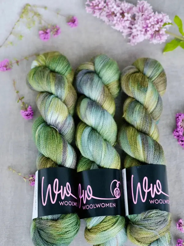 Three skeins of WoolWomen Merino Silk Lace yarn in green and purple are displayed side by side on a light background, accented with small purple flowers and green leaves. Each skein features the WOOLWOMEN label.