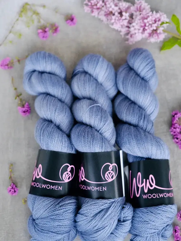 Three skeins of WoolWomen Merino Silk Lace yarn in soft, light purple are arranged side by side on a surface, with lilac flowers and petals scattered around them.