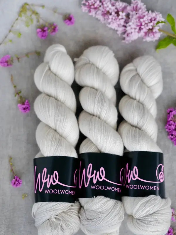 Three skeins of off-white WoolWomen Merino Silk Lace yarn are neatly arranged on a gray surface with purple flowers—perfect for your next käsinvärjätty merinovilla or pitsilanka project.