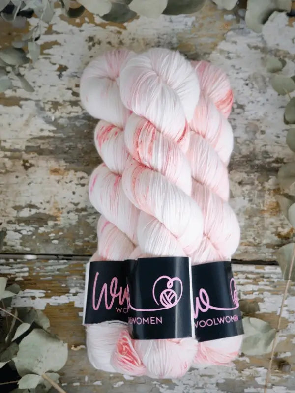 Three skeins of WoolWomen Merino Sock in white with pink speckles are arranged side by side on rustic wood, each labeled WOOLWOMEN. Pale dried leaves frame the display, highlighting the beauty of this unique yarn.