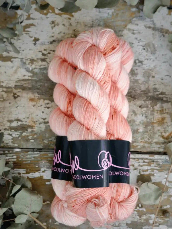 Two skeins of WoolWomen Merino Sock, käsinvärjätty lanka in pale pink with subtle speckles and black WOOLWOMEN labels, are set on a rustic wooden surface with dried leaves around them.